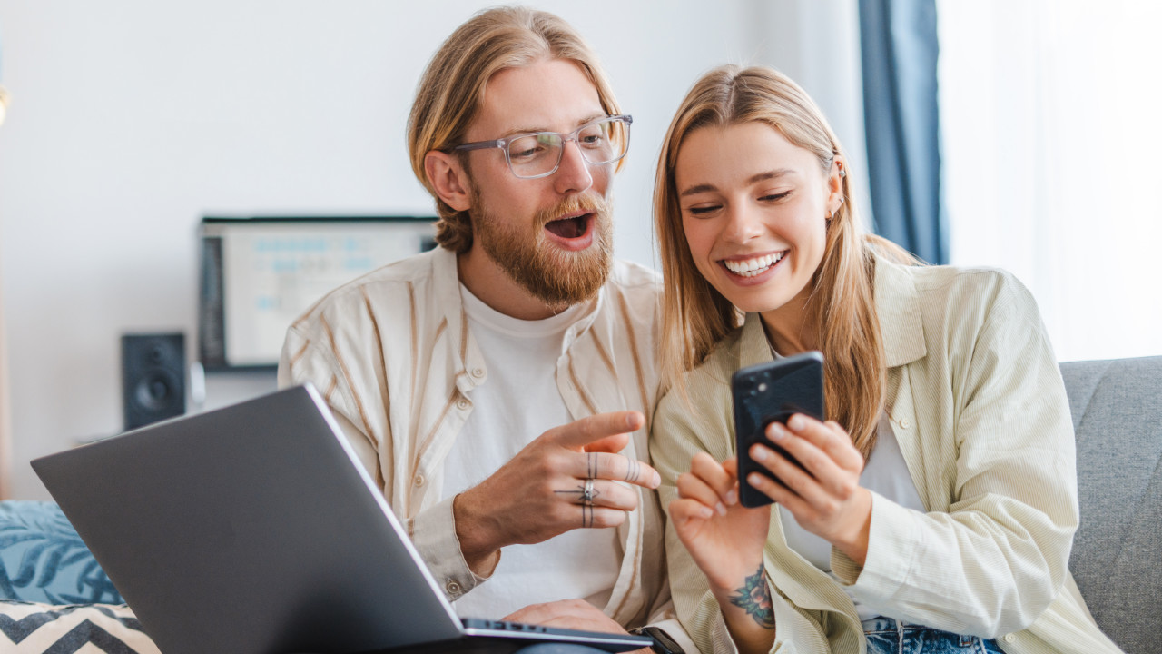 Junges Paar sitzt auf der Couch und blickt lächelnd auf Notebook und Smartphone.